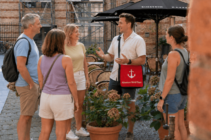Tour guide talks to four tourists in outdoor cafe with brick walls.