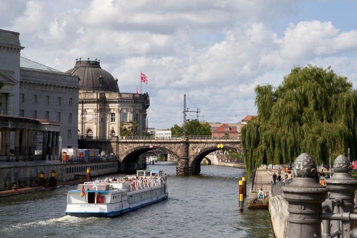 Riverboat cruising past historic buildings and a bridge under a cloudy sky.