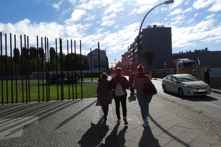 People walking on a sunlit urban street with tall structures and a car nearby.