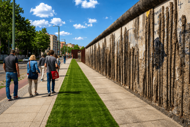 People walk beside a graffiti-covered wall under a blue sky with fluffy clouds.