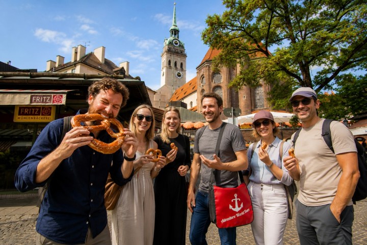 Group of six people smiling with pretzels outdoors near historic building.