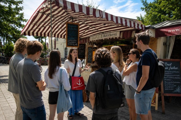 Group of people listening to a woman outside a striped awning bakery stall.