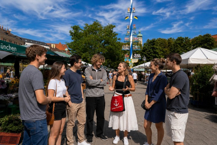 Group of seven people talking and smiling at an outdoor market under a sunny sky.