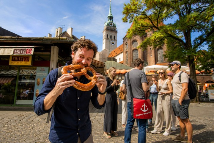 Man enjoying large pretzel outdoors with group of people and historic building in background.