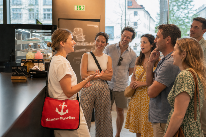 Tour guide talks to a group of smiling adults in a cafe-like setting.