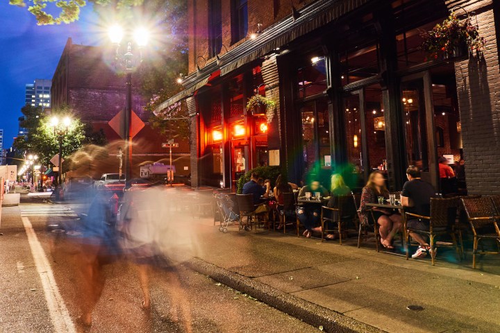 Street view of a bustling outdoor restaurant at night with blurred pedestrians and parked cars.