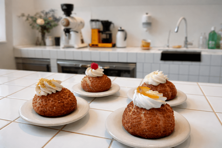 Four cream-topped pastries on plates on a white tiled counter in a kitchen setting.