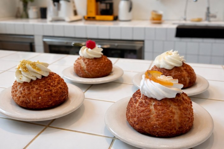 Four cream puffs with whipped cream toppings on white plates on a tiled kitchen counter.