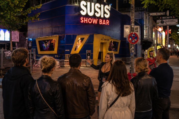 People stand outside a brightly lit show bar at night.
