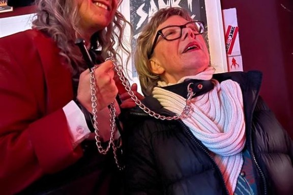 Two people smiling indoors; one holds a chain, wearing coats.