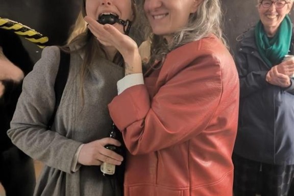 Two smiling women, one in a red coat, the other with a bottle, in a casual indoor setting.