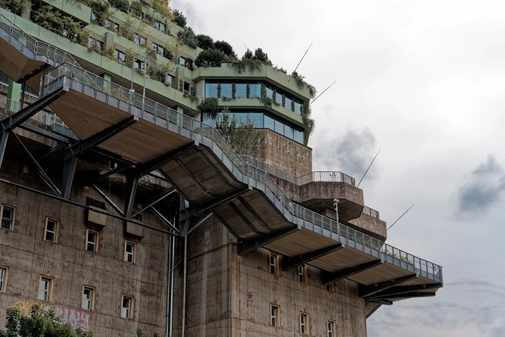 Tiered concrete structure with greenery on top, featuring balconies and angular lines against a cloudy sky.