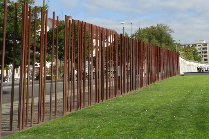 Tall rust-colored metal rods form a monument on a grassy area, with trees and buildings in the background.